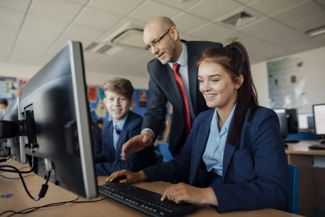 A teacher speaks to a pupil sat at a desktop computer