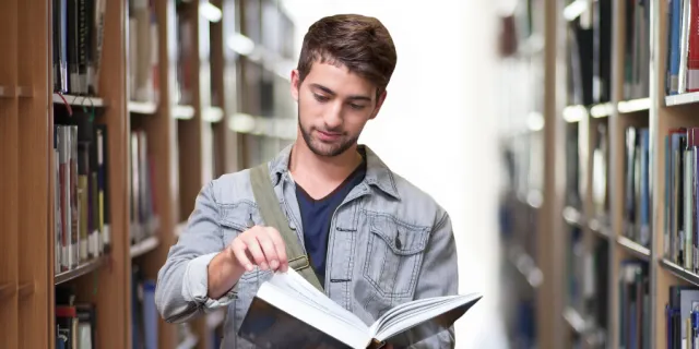 A university students stands between library shelves looking at a book